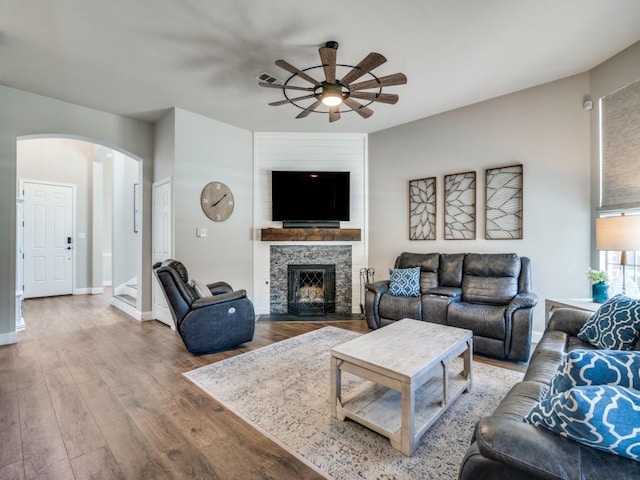 living area featuring arched walkways, ceiling fan, a stone fireplace, wood finished floors, and baseboards