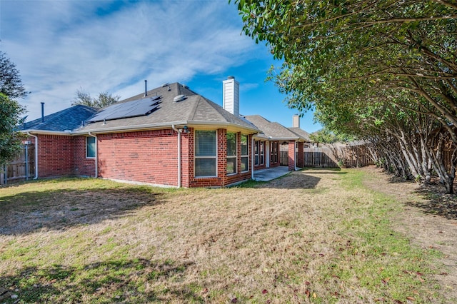 back of house featuring brick siding, a chimney, a lawn, roof mounted solar panels, and a fenced backyard