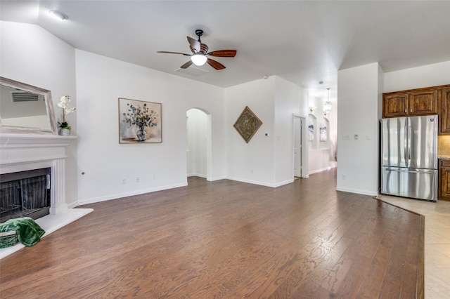 unfurnished living room with a fireplace with raised hearth, arched walkways, wood finished floors, a ceiling fan, and baseboards