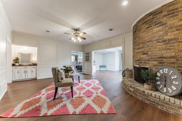 living room featuring dark wood finished floors, ceiling fan, crown molding, a brick fireplace, and a decorative wall