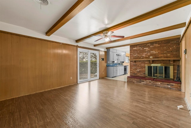 unfurnished living room featuring wooden walls, wood finished floors, a ceiling fan, a brick fireplace, and beamed ceiling