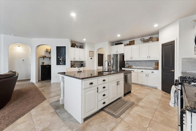 kitchen with arched walkways, stainless steel appliances, a sink, white cabinetry, and a center island with sink