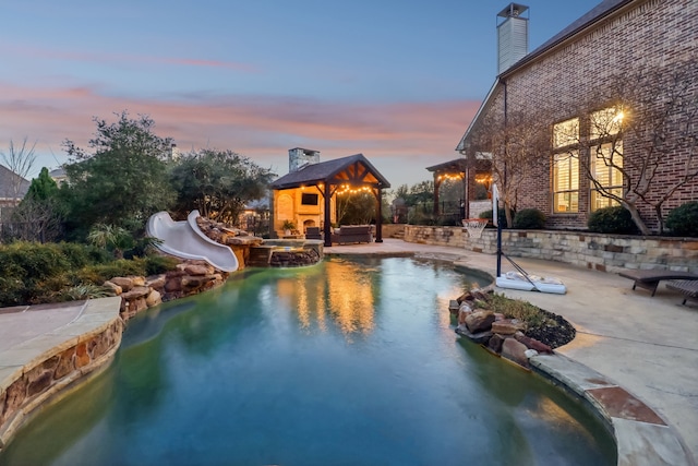 pool at dusk featuring a water slide, a patio area, and a pool with connected hot tub