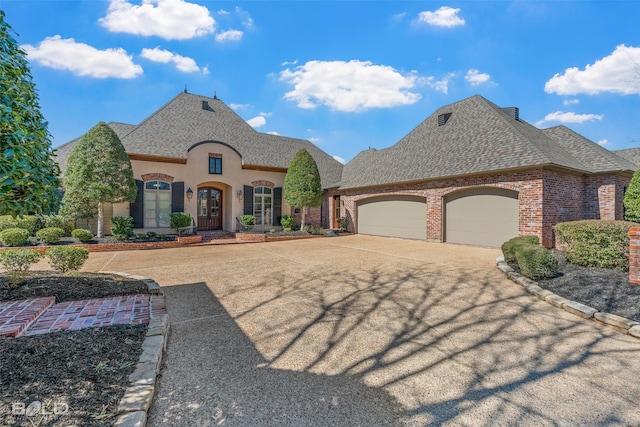 french provincial home featuring a garage, driveway, a shingled roof, and brick siding