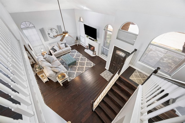 living room with a warm lit fireplace, high vaulted ceiling, wood finished floors, a ceiling fan, and baseboards