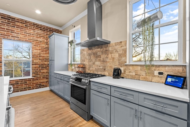 kitchen with brick wall, gray cabinets, light wood-type flooring, wall chimney range hood, and stainless steel range with gas stovetop