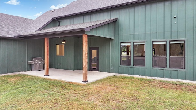 rear view of house with board and batten siding, a patio area, a yard, and roof with shingles