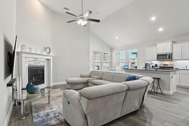 living room featuring baseboards, visible vents, high vaulted ceiling, light wood-style flooring, and a tiled fireplace