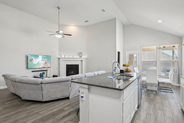 kitchen with a sink, dark stone counters, light wood-style floors, a glass covered fireplace, and a kitchen island with sink