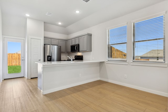 kitchen with light wood-style flooring, appliances with stainless steel finishes, a peninsula, gray cabinetry, and backsplash