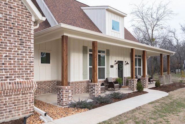 view of exterior entry with a porch, board and batten siding, a shingled roof, and brick siding