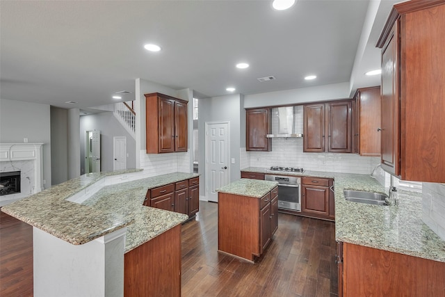 kitchen featuring dark wood-style flooring, appliances with stainless steel finishes, a sink, a peninsula, and wall chimney exhaust hood