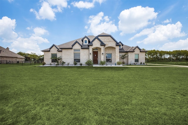 french country style house featuring brick siding, fence, stone siding, roof with shingles, and a front yard