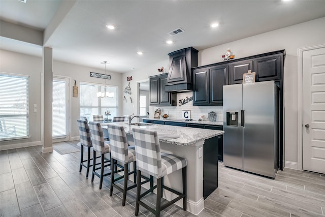 kitchen with a breakfast bar area, visible vents, custom exhaust hood, stainless steel fridge with ice dispenser, and a center island with sink