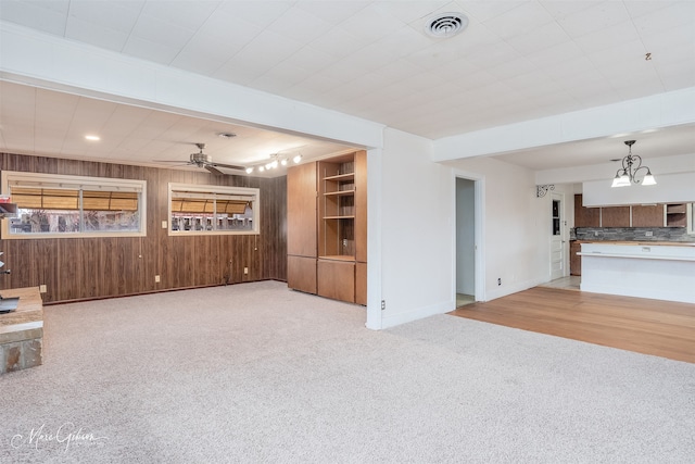 unfurnished living room with wood walls, carpet, visible vents, and ceiling fan with notable chandelier