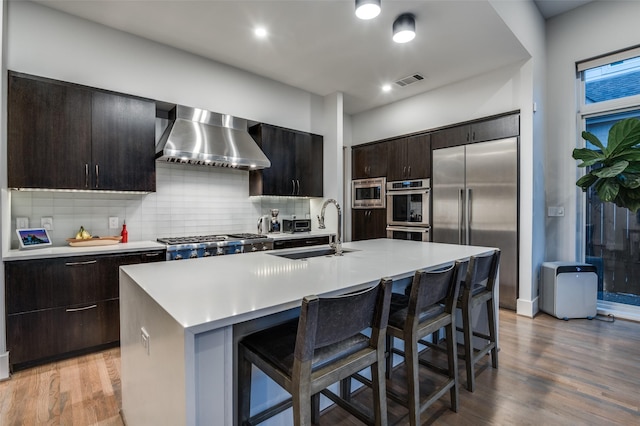 kitchen with light wood finished floors, a sink, wall chimney range hood, built in appliances, and a kitchen bar