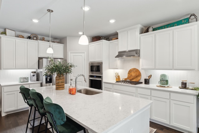 kitchen featuring appliances with stainless steel finishes, white cabinetry, a sink, an island with sink, and under cabinet range hood