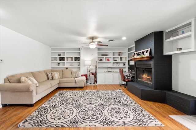 living room with built in shelves, visible vents, a ceiling fan, a brick fireplace, and light wood-type flooring