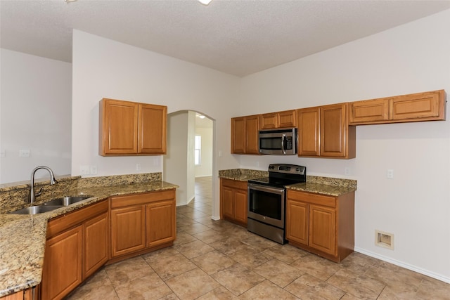 kitchen featuring arched walkways, brown cabinets, light stone countertops, stainless steel appliances, and a sink