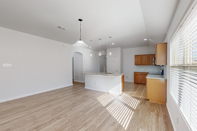 kitchen featuring light wood finished floors, arched walkways, an island with sink, brown cabinets, and a sink