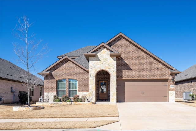 french country inspired facade with a garage, central AC unit, concrete driveway, and brick siding