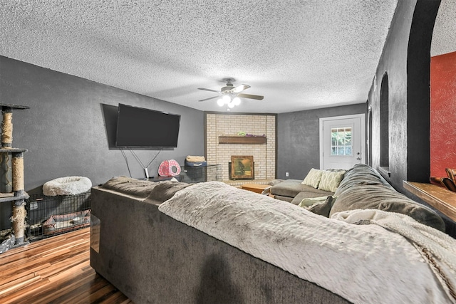 living area featuring a brick fireplace, a ceiling fan, wood finished floors, and a textured wall