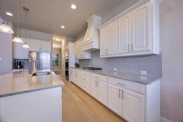 kitchen with light wood finished floors, custom range hood, appliances with stainless steel finishes, white cabinets, and a sink