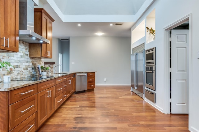 kitchen with wall chimney exhaust hood, brown cabinets, built in appliances, light wood-type flooring, and a warming drawer