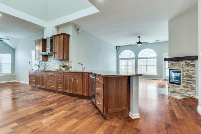 kitchen featuring brown cabinetry, a ceiling fan, wall chimney exhaust hood, a peninsula, and stone counters