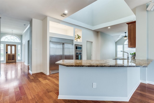 kitchen with built in appliances, wood finished floors, a sink, visible vents, and dark stone countertops