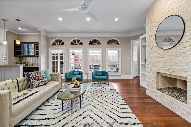 living area with a ceiling fan, dark wood-type flooring, crown molding, and a stone fireplace