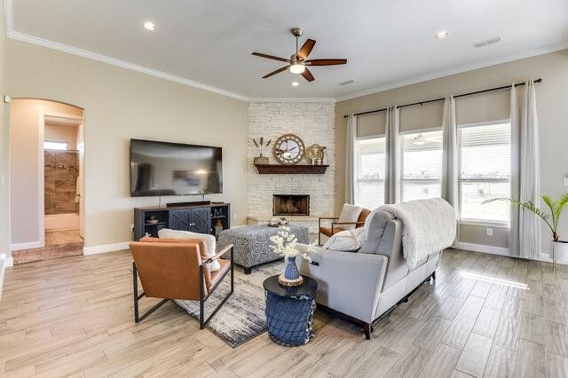 living area featuring arched walkways, crown molding, a stone fireplace, light wood-type flooring, and baseboards