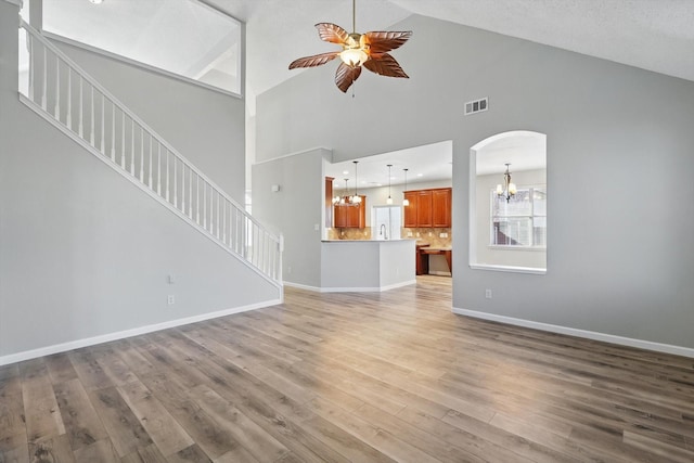 unfurnished living room featuring ceiling fan with notable chandelier, wood finished floors, visible vents, baseboards, and stairway