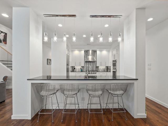 kitchen featuring dark wood-style floors, dark countertops, backsplash, a peninsula, and wall chimney exhaust hood