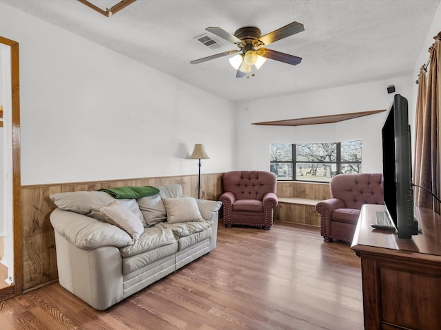 living area featuring ceiling fan, a textured ceiling, wood walls, wood finished floors, and wainscoting