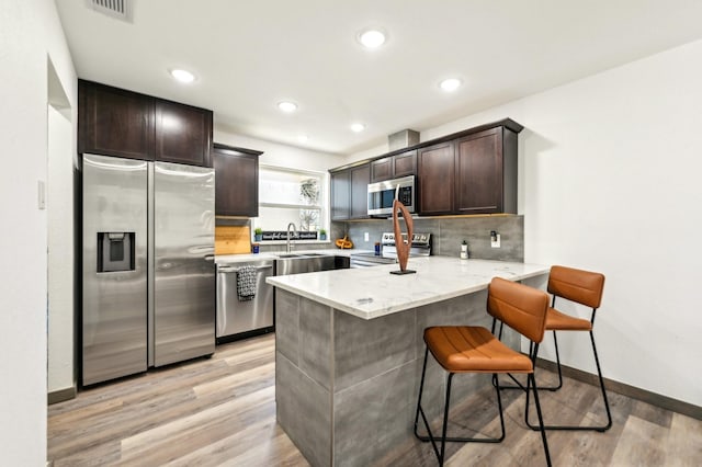 kitchen with decorative backsplash, appliances with stainless steel finishes, dark brown cabinets, light wood-type flooring, and a peninsula