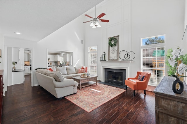 living area with dark wood-type flooring, a fireplace with flush hearth, a ceiling fan, high vaulted ceiling, and baseboards