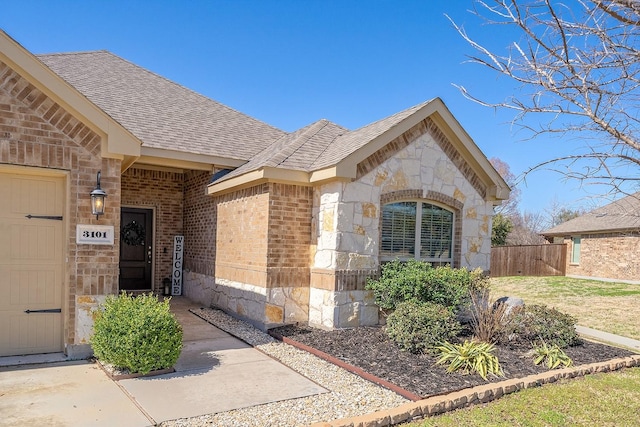 view of exterior entry featuring a shingled roof, stone siding, an attached garage, fence, and brick siding