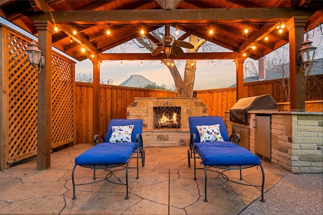 patio terrace at dusk featuring grilling area, fence, exterior kitchen, a gazebo, and an outdoor stone fireplace