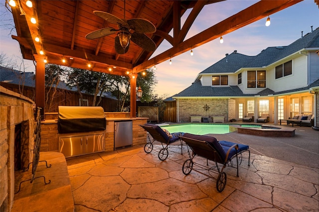 patio terrace at dusk featuring an outdoor kitchen, a fenced backyard, a grill, a ceiling fan, and a gazebo