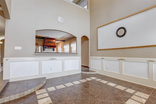 unfurnished living room featuring arched walkways, a decorative wall, a towering ceiling, visible vents, and wainscoting