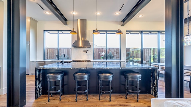 kitchen featuring wall chimney range hood, dark stone countertops, light wood-style floors, and beamed ceiling