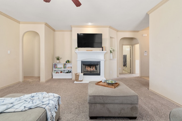 carpeted living room featuring ceiling fan, arched walkways, a fireplace with raised hearth, and ornamental molding