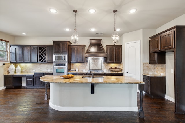 kitchen featuring stainless steel appliances, premium range hood, a kitchen island with sink, and dark brown cabinets