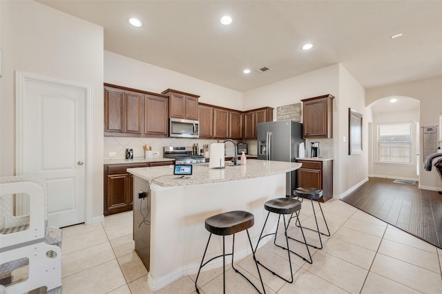 kitchen featuring light tile patterned floors, arched walkways, decorative backsplash, a kitchen island with sink, and stainless steel appliances