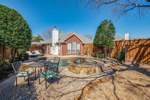 back of house featuring a fenced backyard, brick siding, a pool with connected hot tub, a chimney, and a patio area