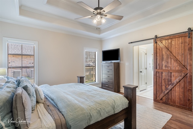 bedroom with a tray ceiling, a barn door, ornamental molding, a ceiling fan, and wood finished floors