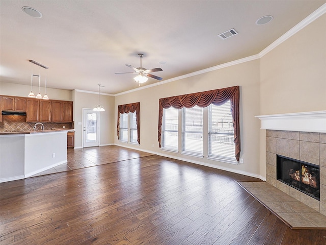 unfurnished living room featuring ceiling fan, visible vents, dark wood-style floors, a tiled fireplace, and crown molding