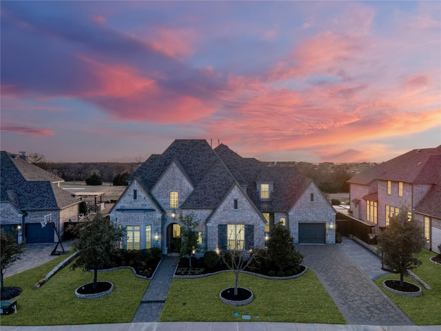 french country style house with an attached garage, stone siding, decorative driveway, and a yard