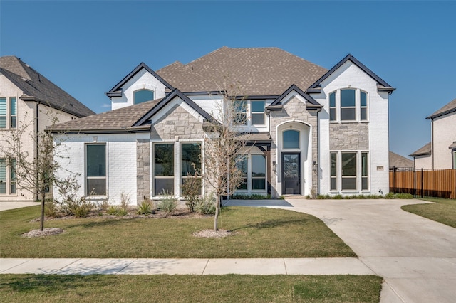 french provincial home with a shingled roof, stone siding, fence, a front yard, and brick siding